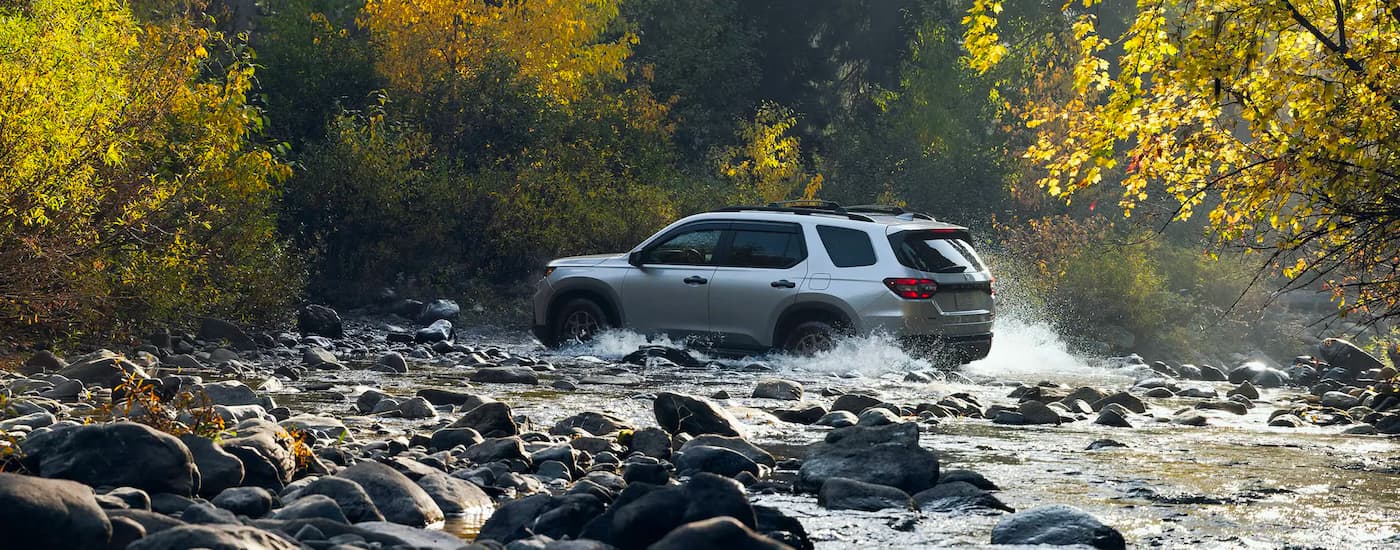 Left angle view of a grey 2023 Honda Pilot Trailsport driving over a shallow river.