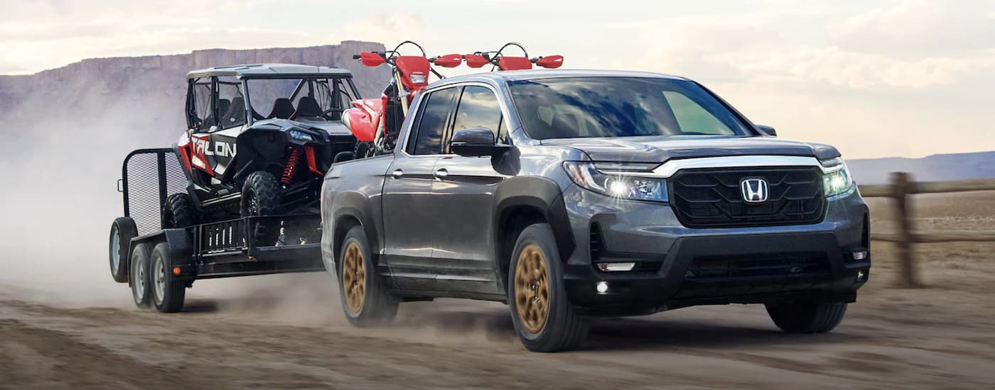 A silver 2022 Honda Ridgeline RTL-E towing ATVs on a dusty road.